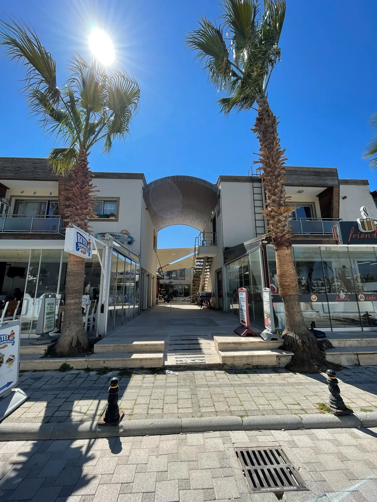 A sunny street scene shows two modern buildings with balconies, separated by a walkway. Tall palm trees line the entrance, and a bright blue sky is overhead. Signs and storefronts are visible on the ground floor.