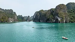 A panoramic view of Ha Long Bay’s emerald waters dotted with towering limestone karsts and islets covered in green vegetation. A few traditional wooden boats and a small rowboat are scattered across the water.