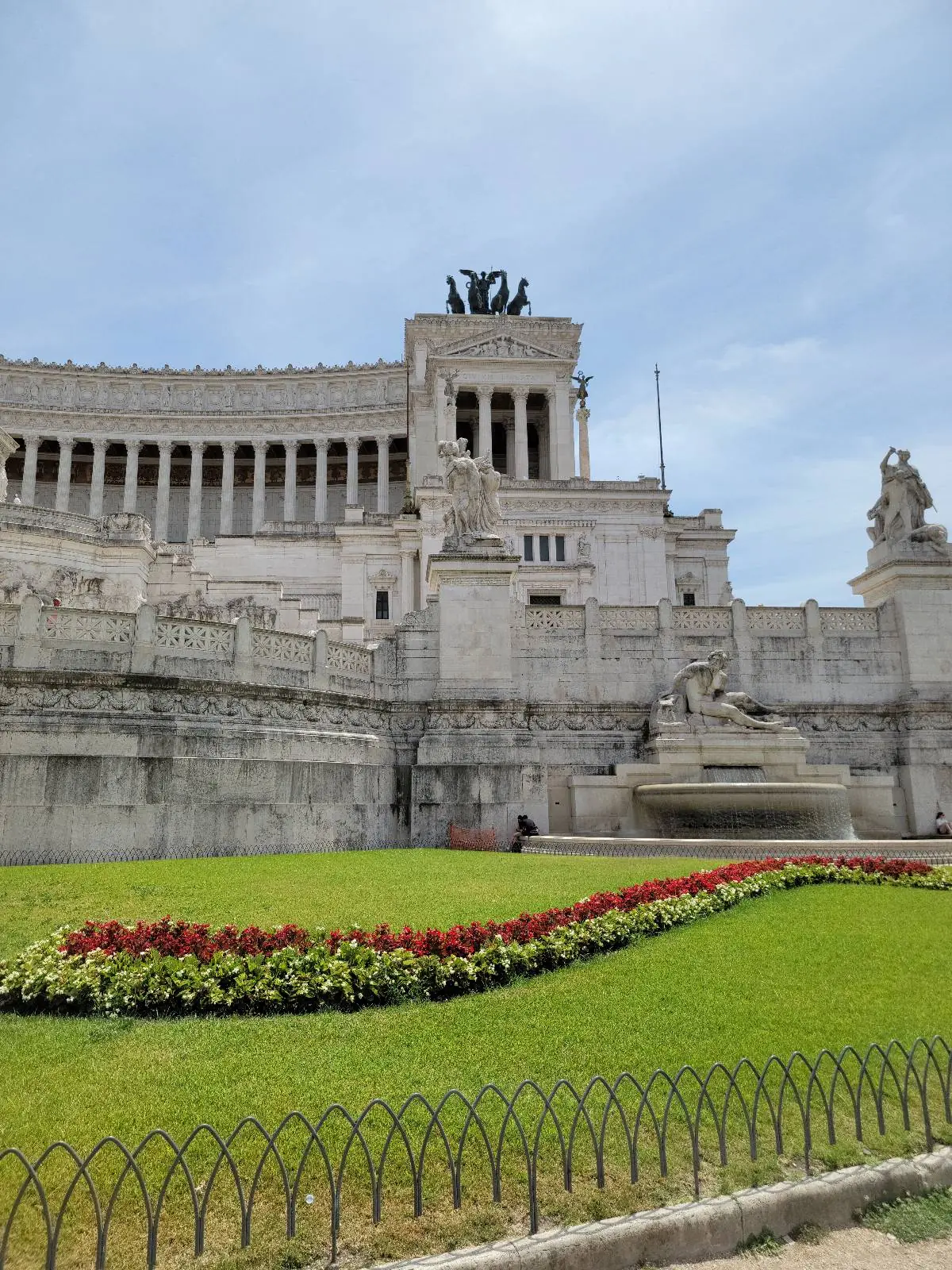 A large white marble monument with statues and columns, surrounded by landscaped gardens with red and green flower beds, under a partly cloudy sky.