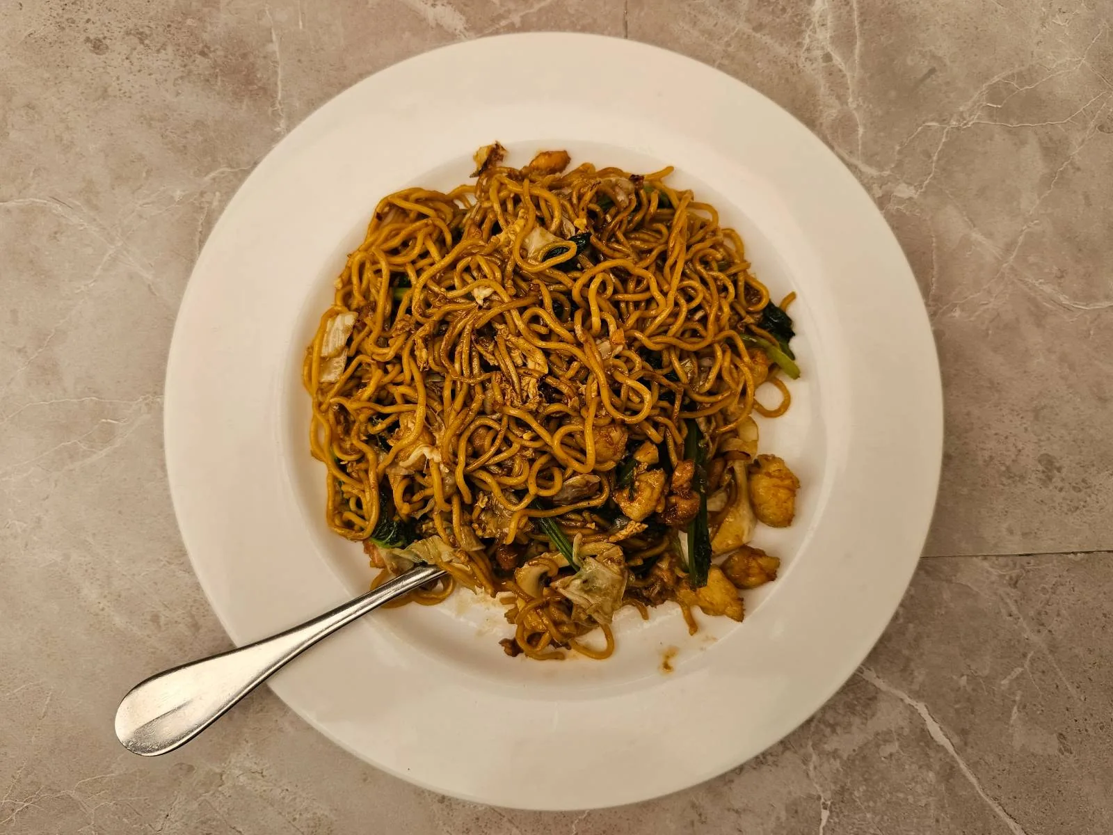 A white plate filled with cooked noodles mixed with vegetables and small pieces of chicken, placed on a marble surface. A silver fork rests on the edge of the plate. The noodles appear seasoned with a dark sauce.