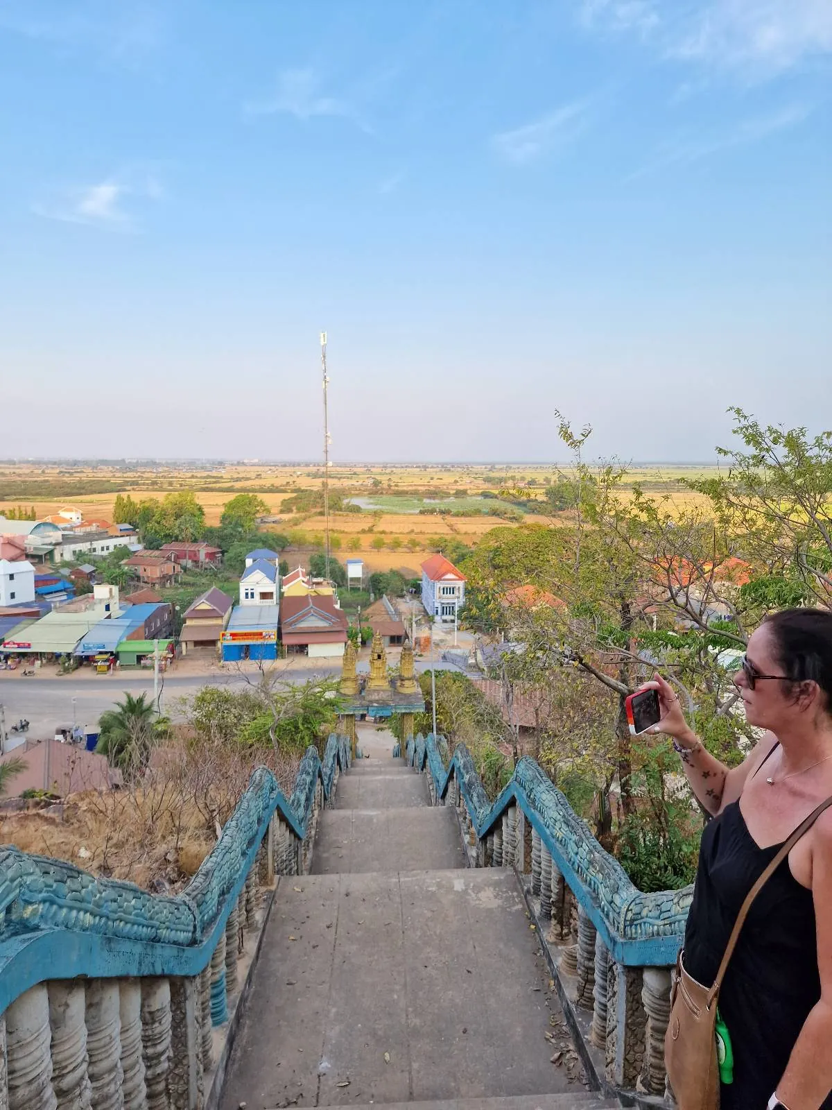 A woman in sunglasses takes a selfie on a stone staircase overlooking a small town, fields, and a clear blue sky on a sunny day.