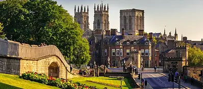 Historic cityscape of York, England, featuring medieval stone walls, lush green trees, and the prominent towers of York Minster cathedral rising above red-brick buildings under a clear sky.