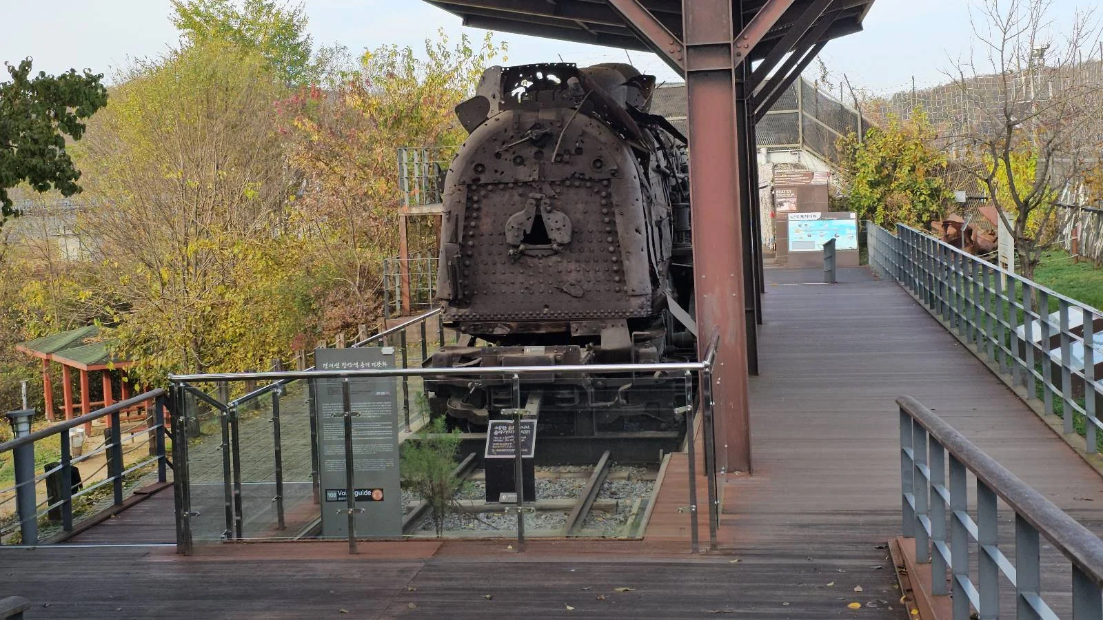 A rusted locomotive on display, framed by a glass barrier on a wooden walkway. Trees with autumn foliage are in the background, and a metal canopy provides partial cover.