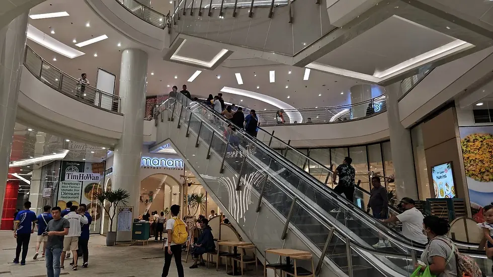People on escalators in a brightly lit mall. "Manam" and "TimHoWan" signs visible. Shoppers walk and gather, creating a lively atmosphere.