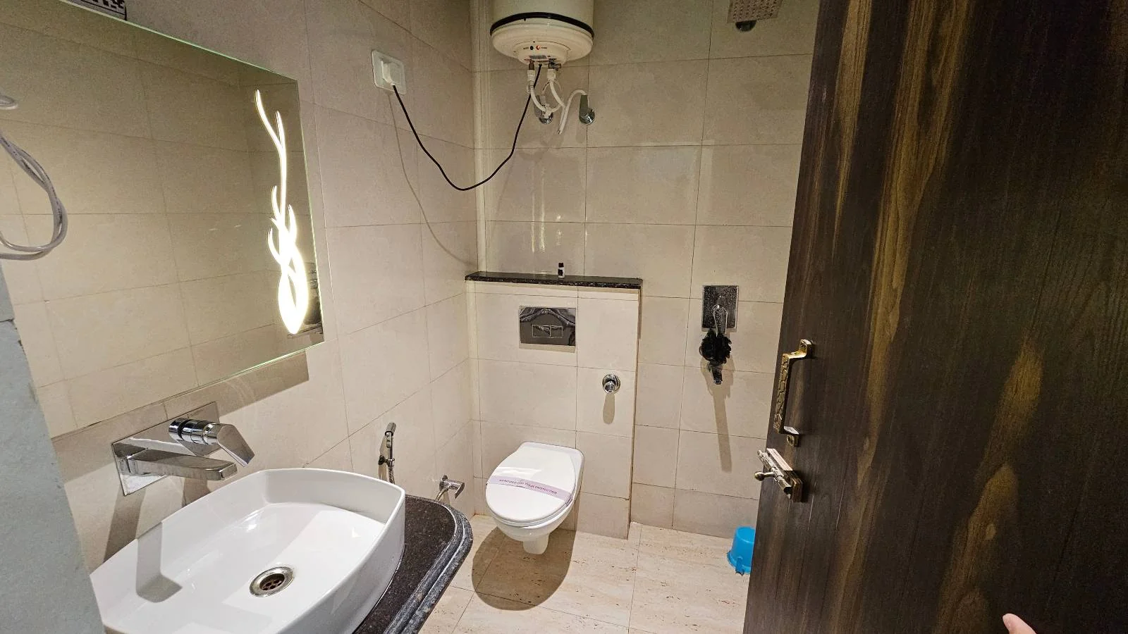 A small bathroom with a sink and faucet on the left, a wall-mounted toilet in the center, and a mirror above the sink. The walls and floor are tiled in light colors. A water heater is mounted above the toilet. The door is slightly open.