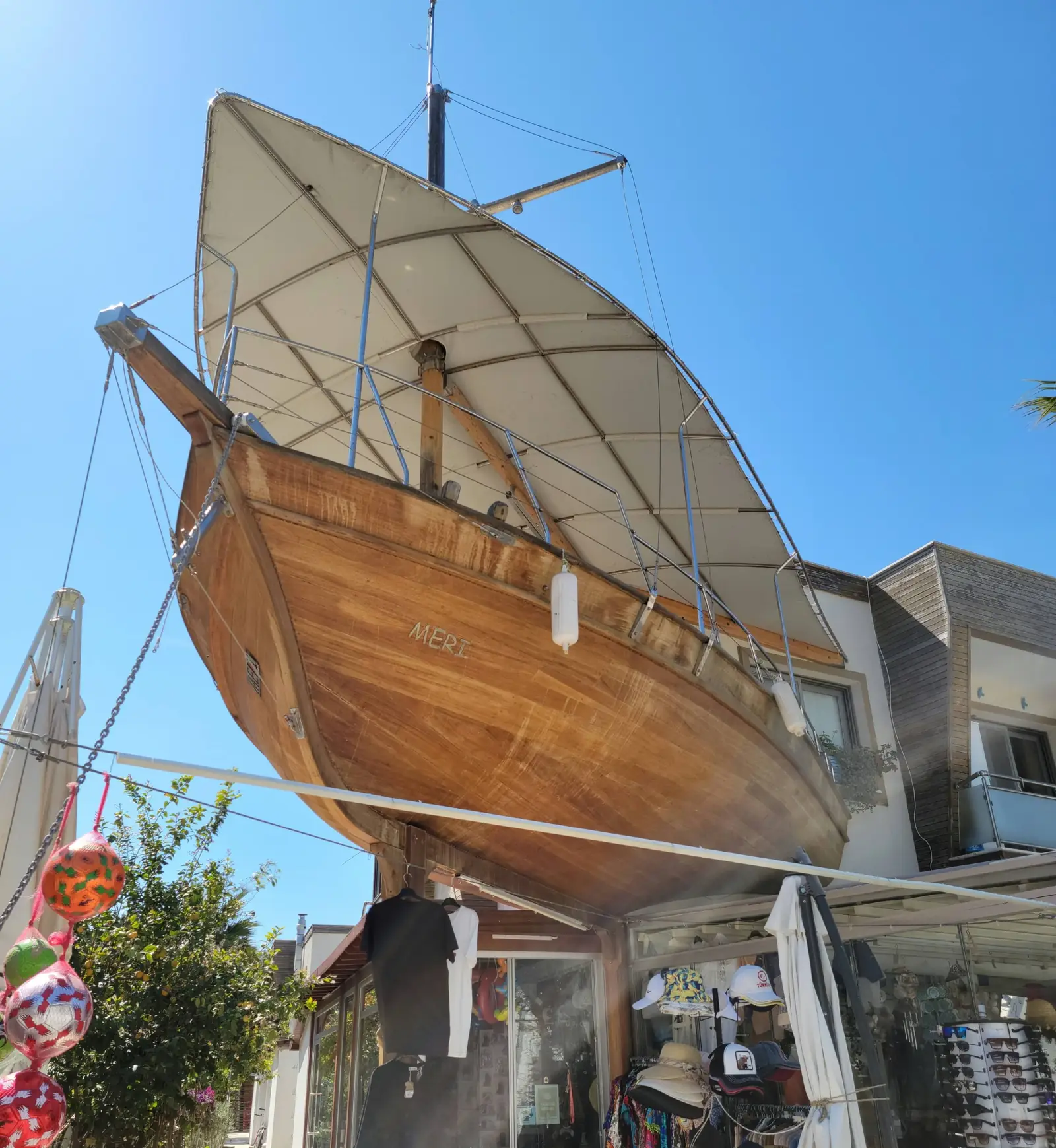 A large wooden boat is suspended above street-level shops, with its sails open, against a bright blue sky. Nearby buildings and market stalls are visible below the boat.