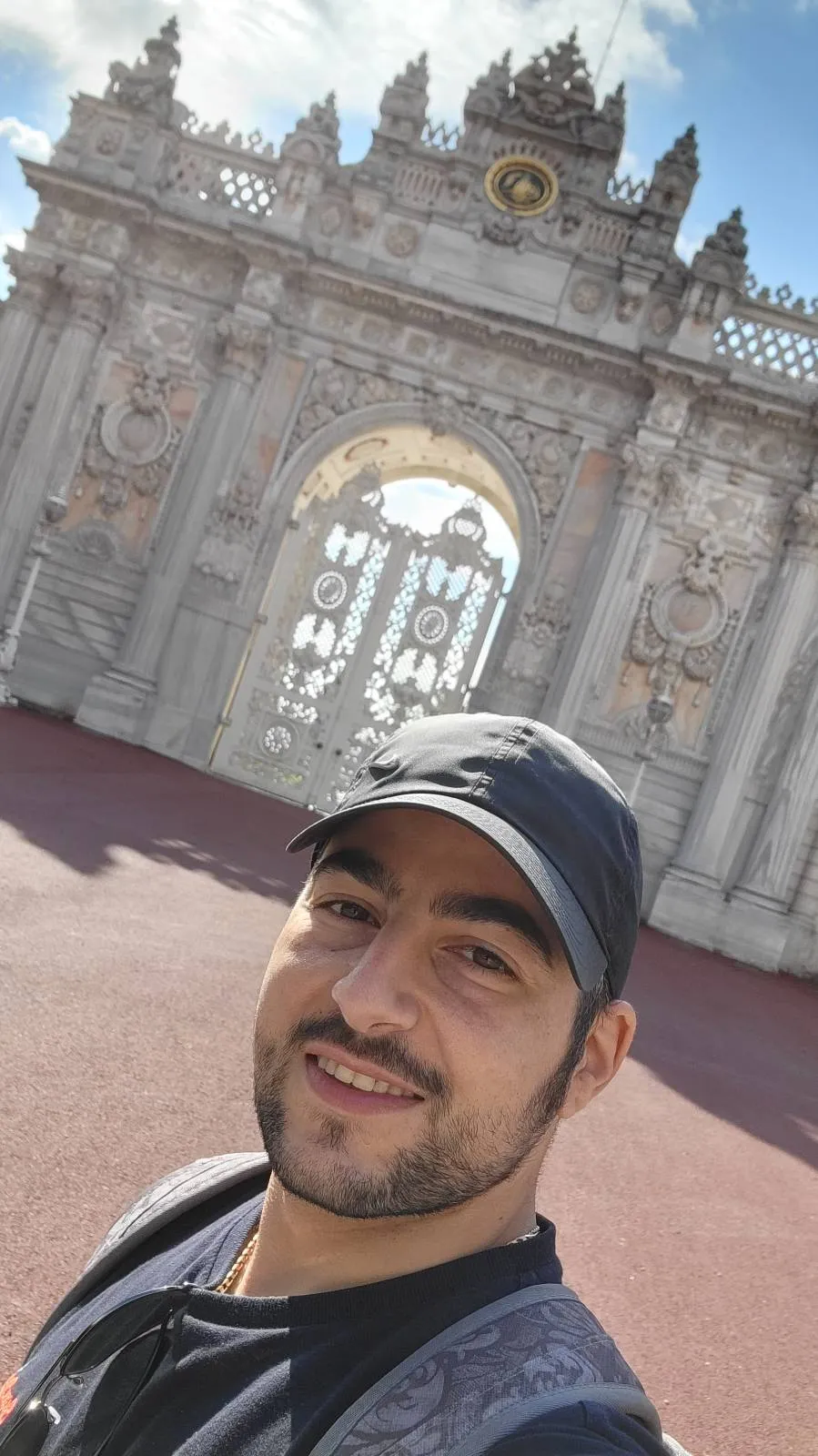 A man wearing a black cap and gray shirt smiles for a selfie in front of an ornate, historic stone gate with decorative details under a partly cloudy sky.