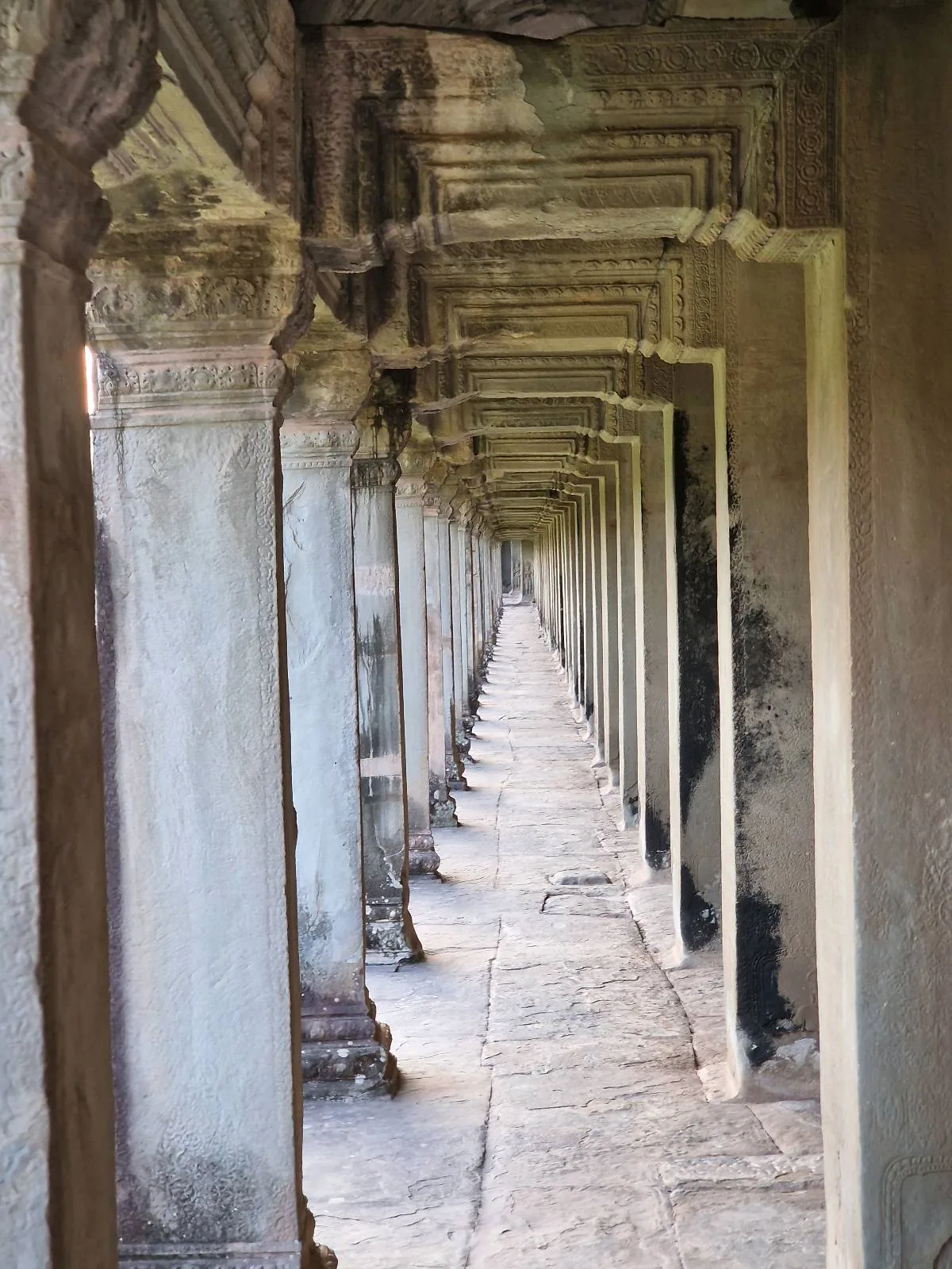 Stone corridor with repeating columns and detailed carvings, creating a sense of depth and symmetry; the worn floor and walls suggest an ancient or historic structure.