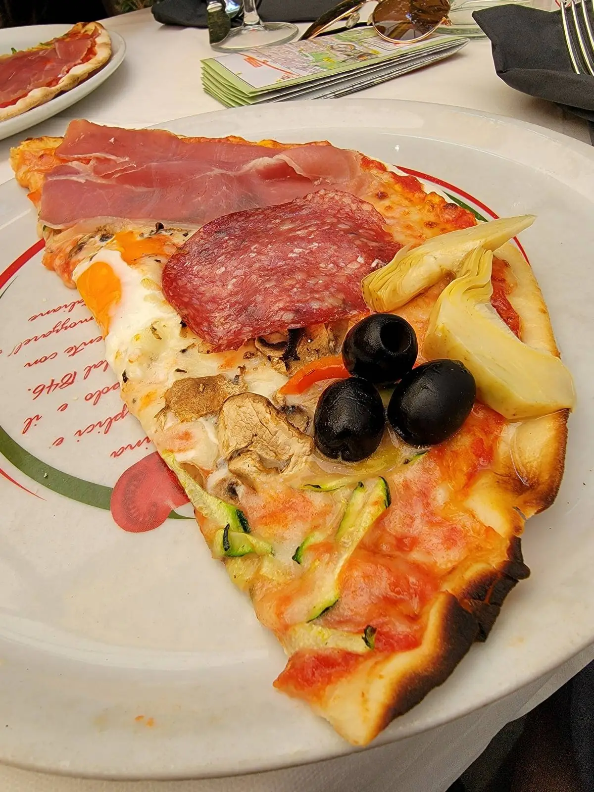 A slice of pizza on a plate, topped with black olives, mushrooms, zucchini, pineapple, cured meats, and melted cheese.