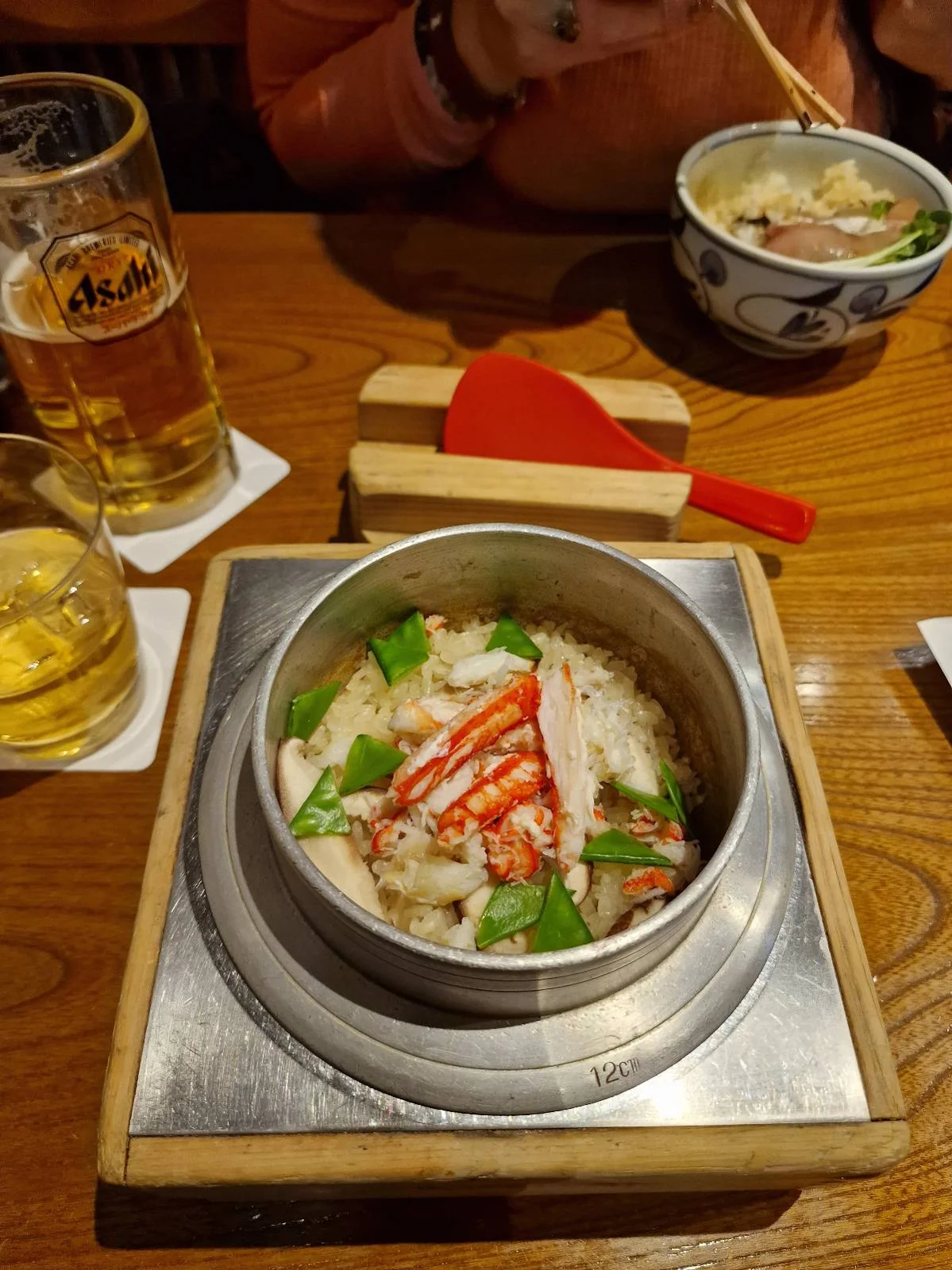 A table with a bowl of rice topped with crab meat and green vegetables, placed on a wooden tray. There's a glass of beer, a glass of water, and a person in the background eating from a bowl with chopsticks.
