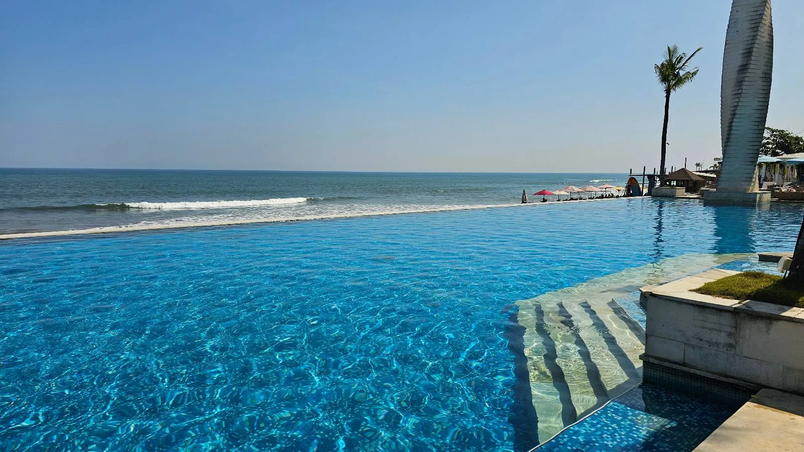 Poolside scene with lounge chairs and blue umbrellas facing an infinity pool. Tall palm trees line the area, and the ocean is visible in the background under a clear blue sky. The setting is sunny and tropical.