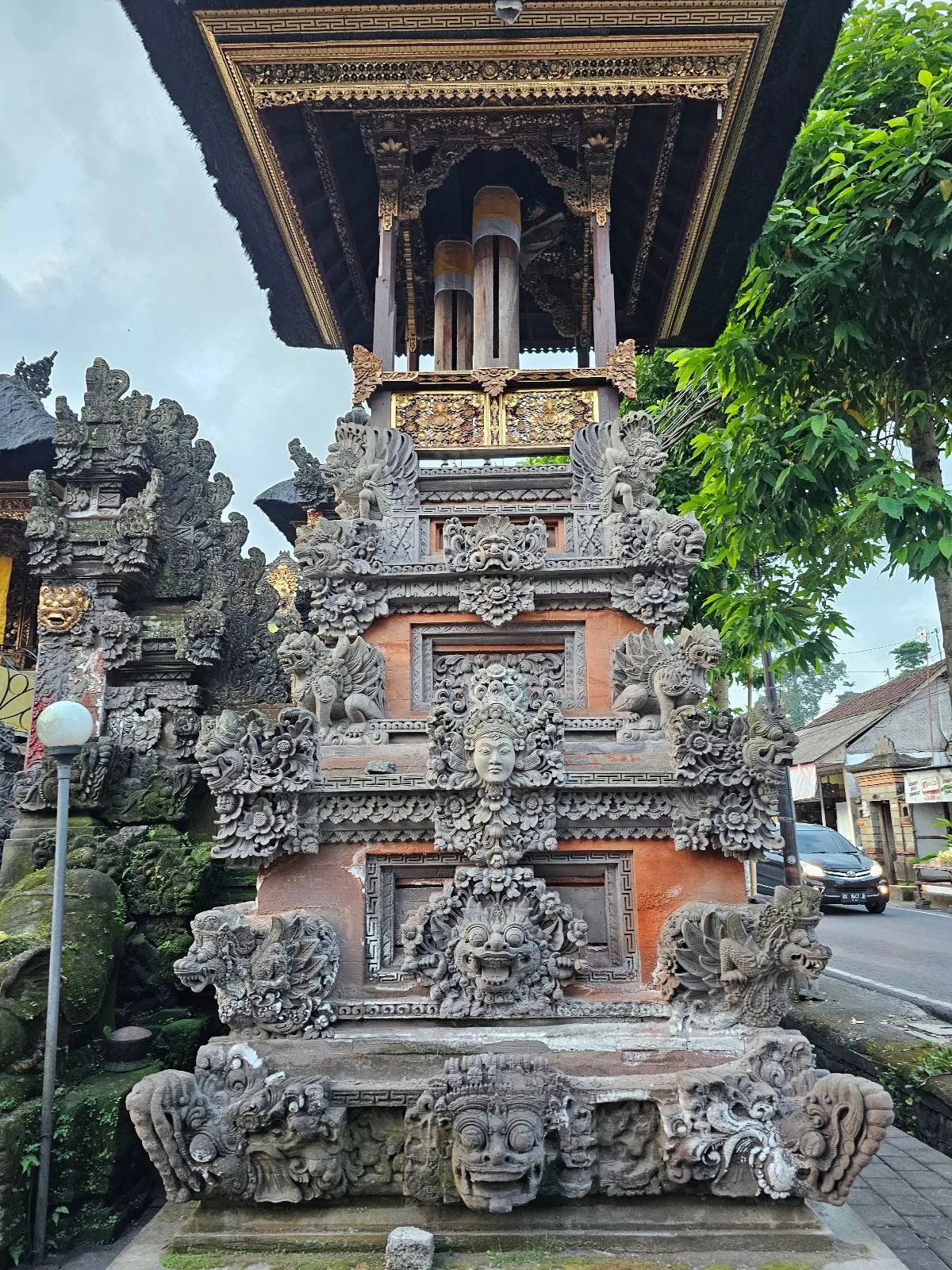Ornate, traditional Balinese stone shrine with intricate carvings of mythical creatures and faces, situated under a wooden pagoda-style roof. Lush greenery surrounds the structure, while buildings are visible in the background.