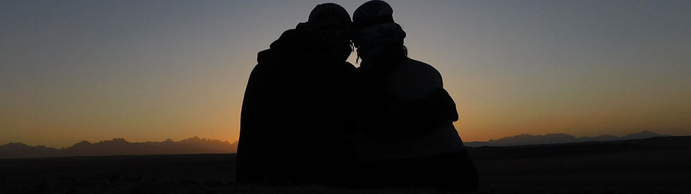 Two people sitting closely together, silhouetted against a dim or dusk sky, suggesting an intimate or peaceful moment.