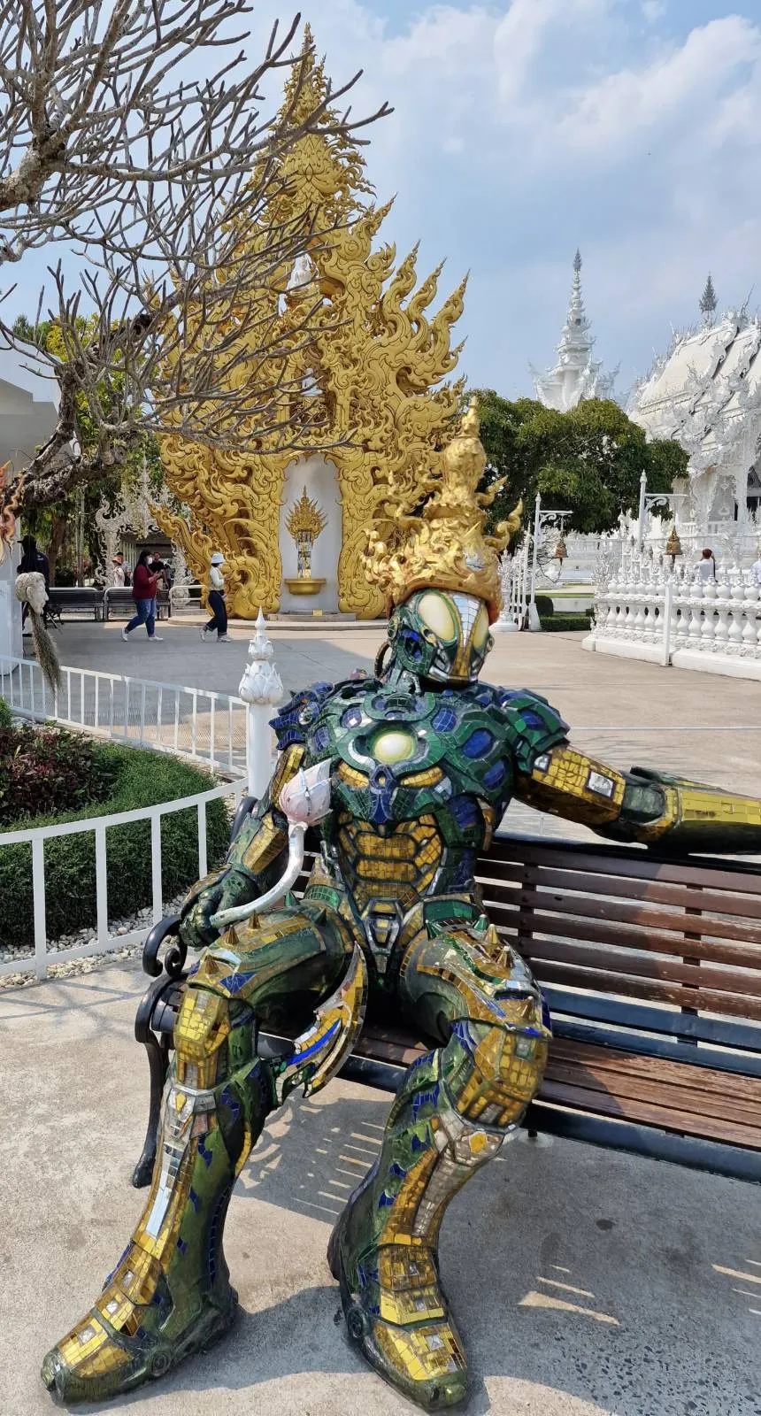 A person in ornate armor sits on a bench in front of a temple with intricate golden and white architecture. The sky is clear and blue, and there are trees and other visitors in the background.