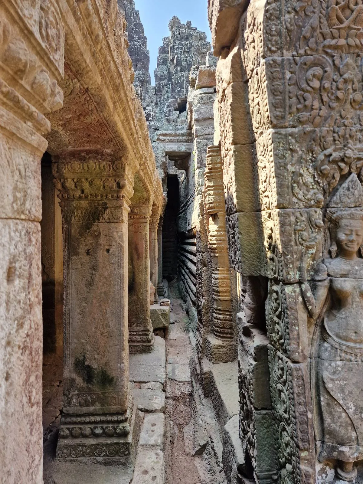 Narrow stone passageway between ancient temple walls, decorated with detailed carvings and a stone relief of a standing female figure; sunlight highlights the weathered textures.