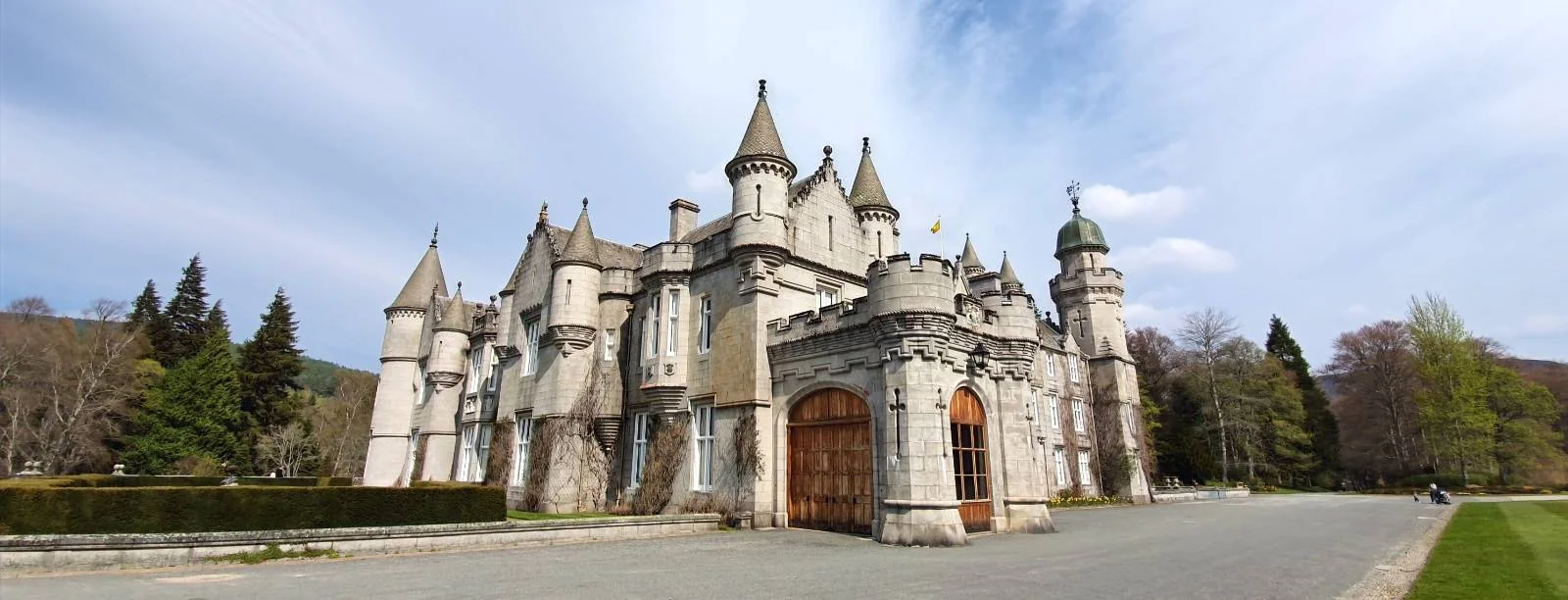 A grand, stone castle with multiple towers and spires stands surrounded by trees and a clear blue sky, with a large wooden gate at the entrance.