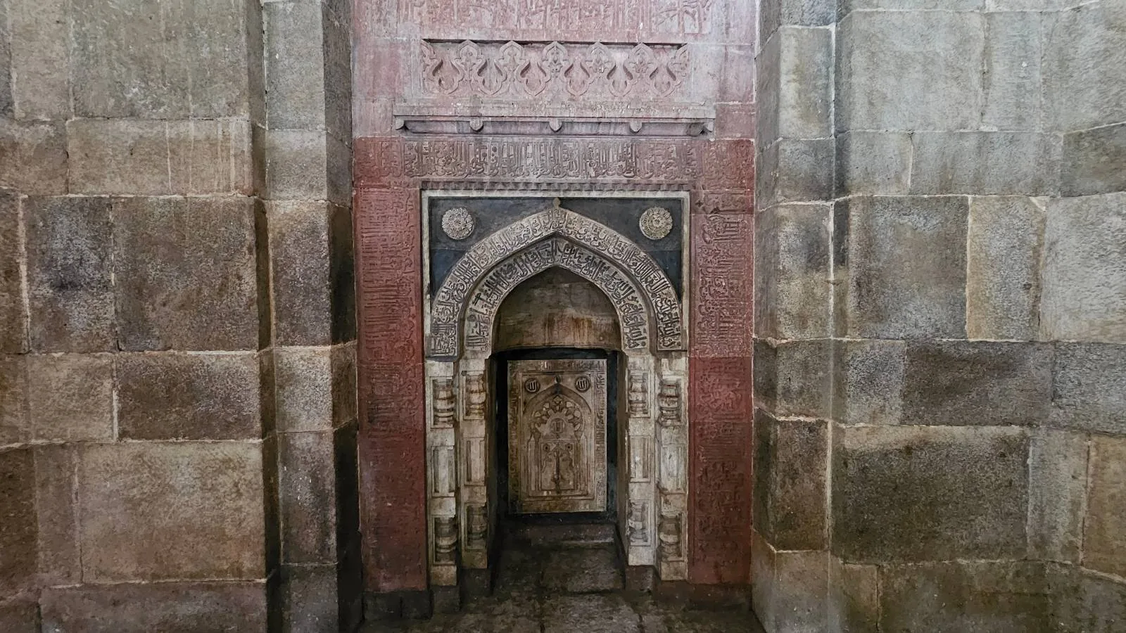 Ornate stone mihrab set into a mosque wall, featuring intricate geometric patterns and arch designs in red and gray, flanked by large stone blocks.