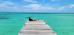 A person in a black swimsuit lies on a wooden pier extending into the clear, turquoise waters off Passion Island, Mexico. Beneath the bright blue sky with fluffy white clouds, this serene scene promises a relaxing day in paradise.