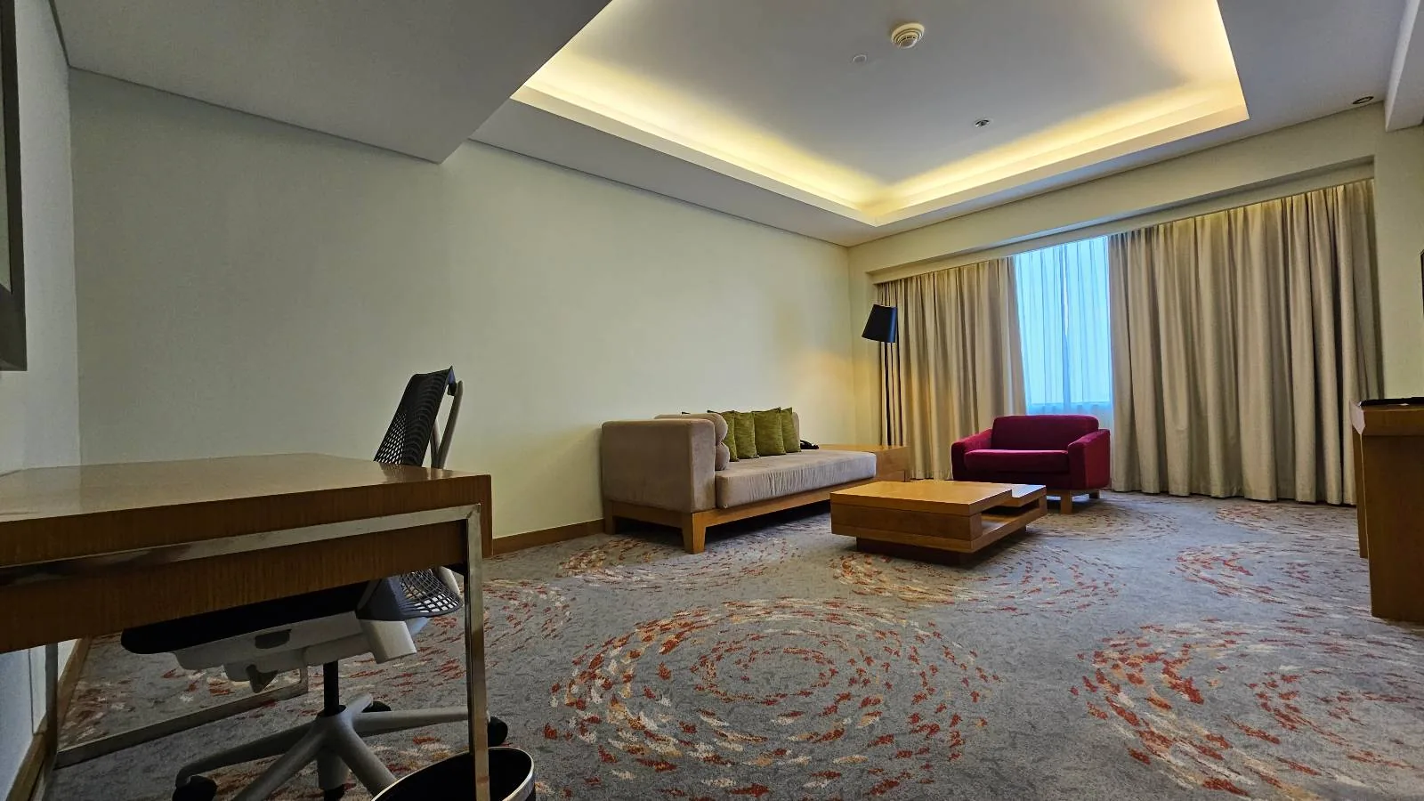 Spacious hotel room with a desk and chair on the left, a beige sofa and red armchair near a coffee table in the center, large window with curtains, and modern lighting on the ceiling.