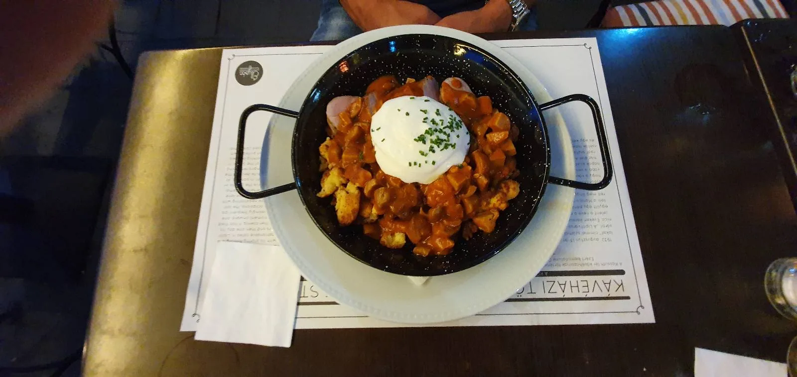 A dish of patatas bravas topped with aioli and garnished with herbs, served in a black pan. The pan rests on a white plate placed on a white placemat, with a napkin nearby on the dark table.