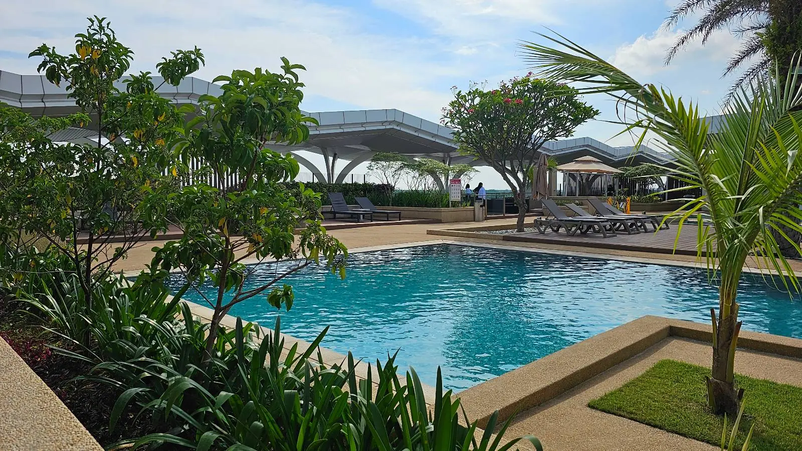 A serene outdoor pool surrounded by lush greenery, including palm trees and various plants, with lounge chairs and modern structures visible in the background under a partly cloudy sky.