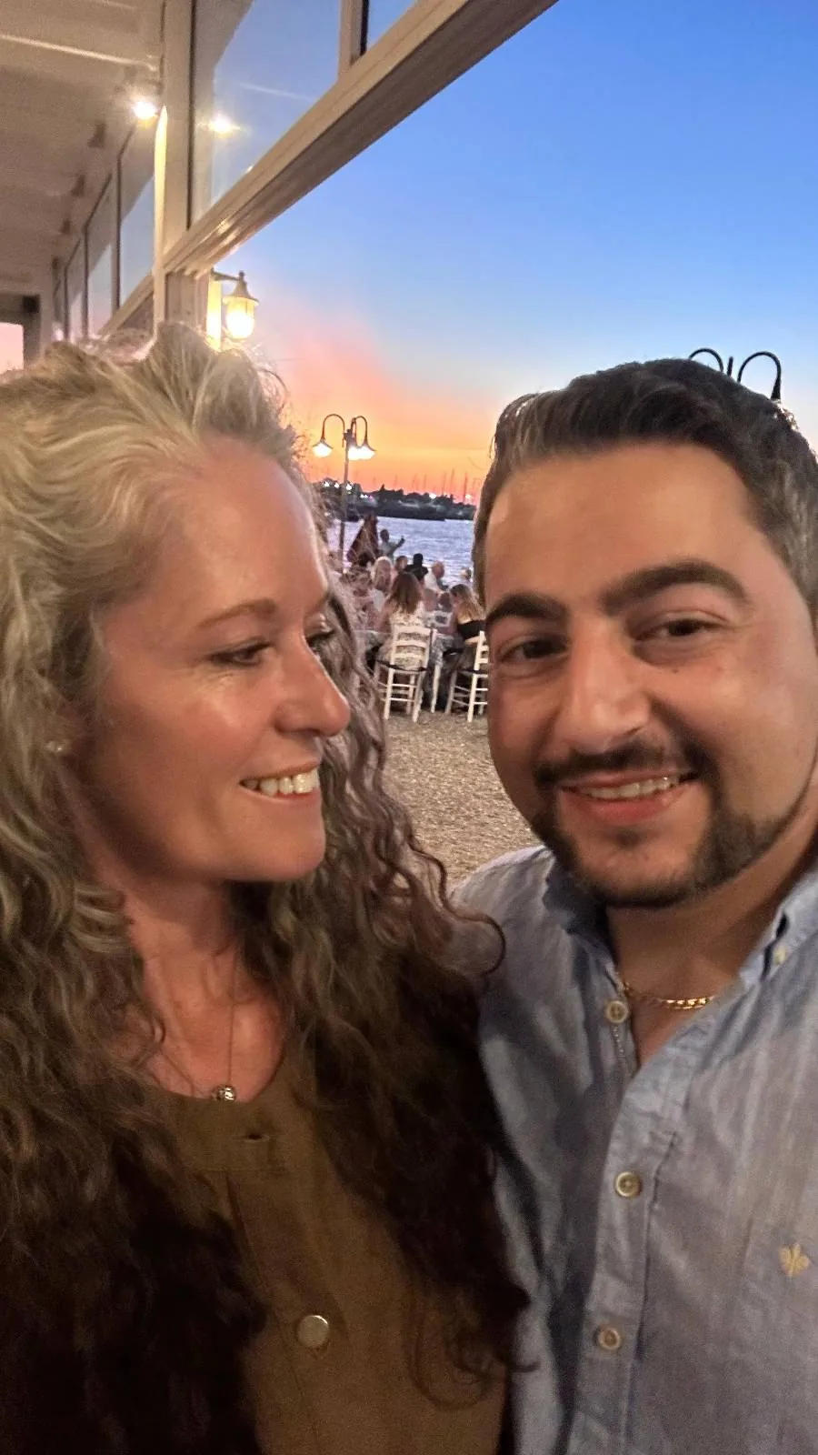 A woman with curly gray hair smiles at a man with short dark hair and a trimmed beard as they take a selfie at an outdoor restaurant during sunset near the sea.