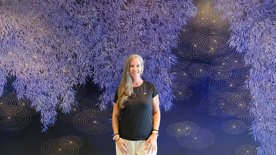 Smiling woman stands before a purple leafy mural with glowing spiral dots on a dark background.
