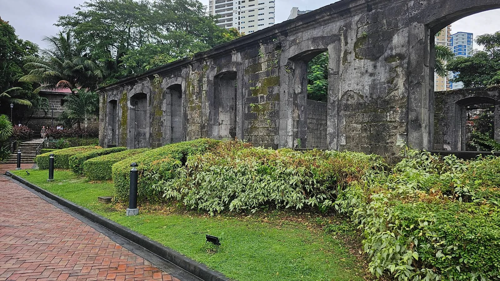 A stone ruin with large arched windows stands beside neatly trimmed shrubs and a brick walkway, surrounded by greenery and tall buildings in the background.