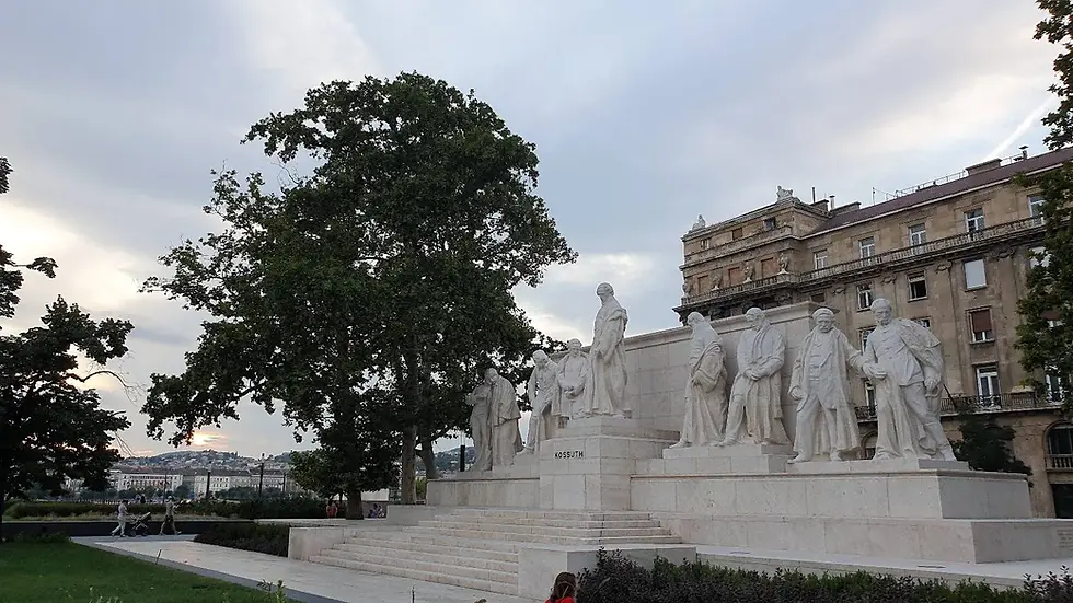 Statue of Kossuth with historical figures on a pedestal. Nearby, people stroll with a stroller. Trees and buildings in the background.