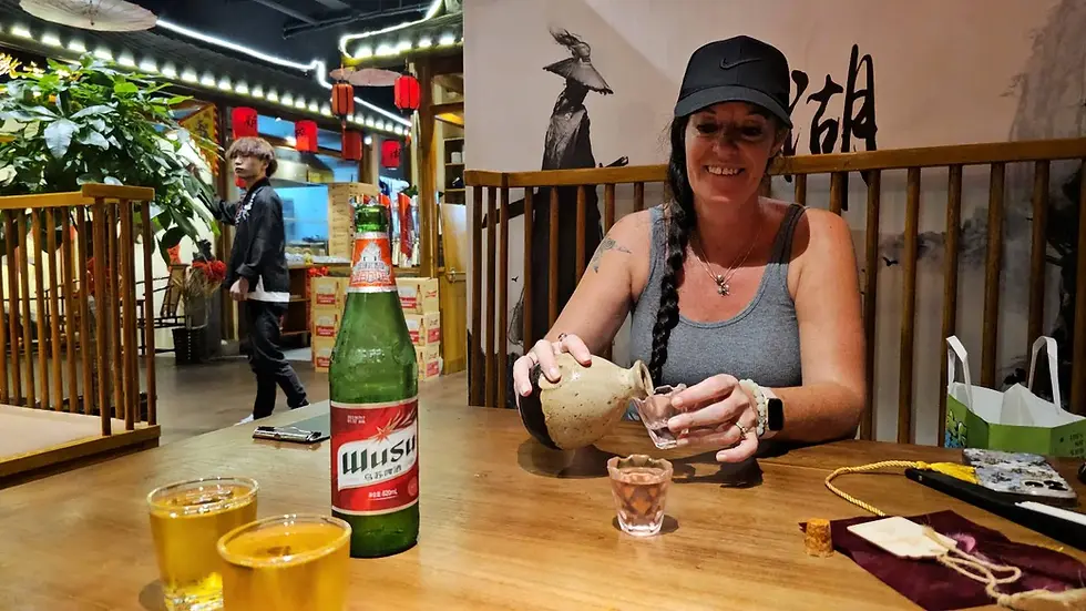 Woman in a cap pours a drink into a glass at a wooden table with a green bottle and yellow drinks. Background shows a traditional decor.