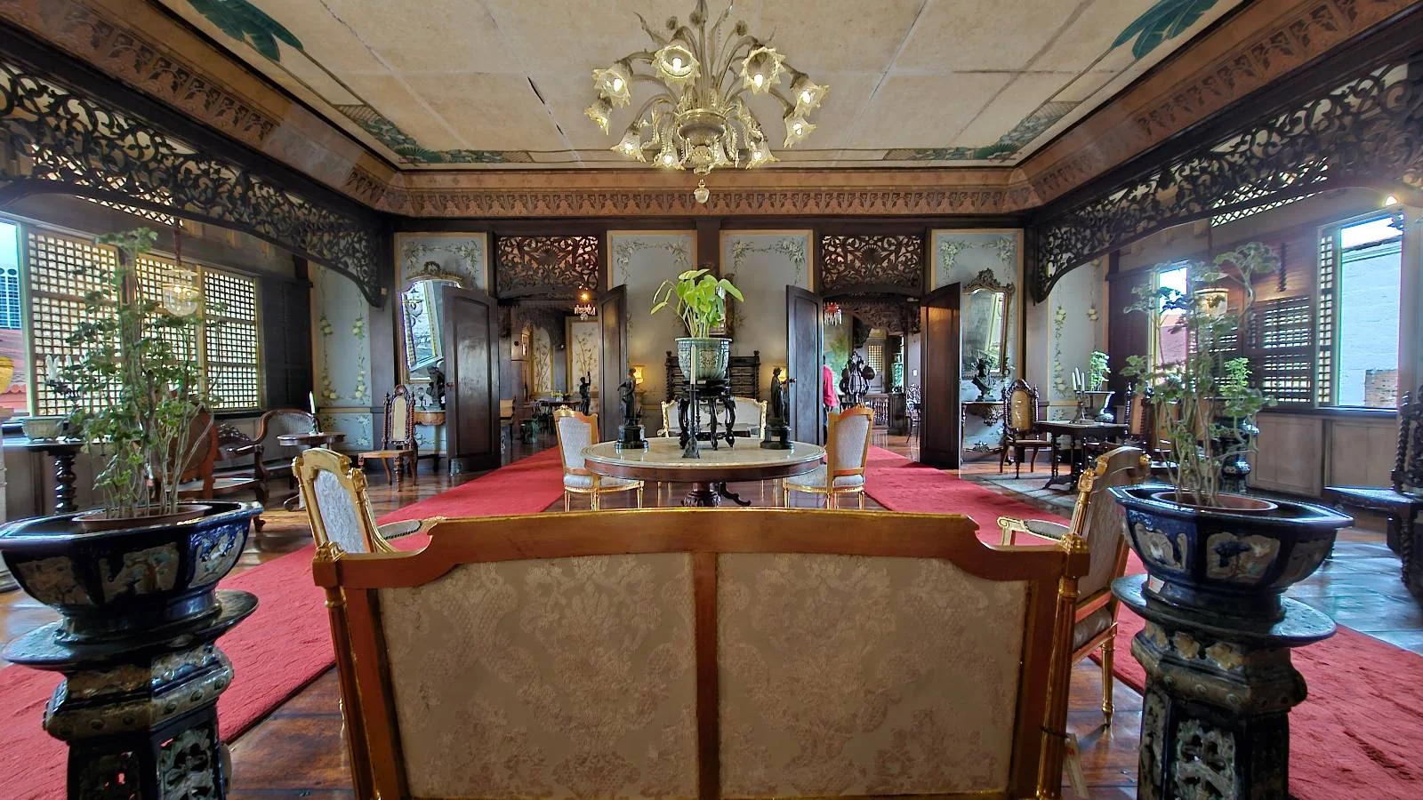A luxurious, ornate room with a large chandelier, red carpeting, and elegant antique furniture. The room is decorated with intricate wood carvings, potted plants, and large windows, creating a classic and refined atmosphere.