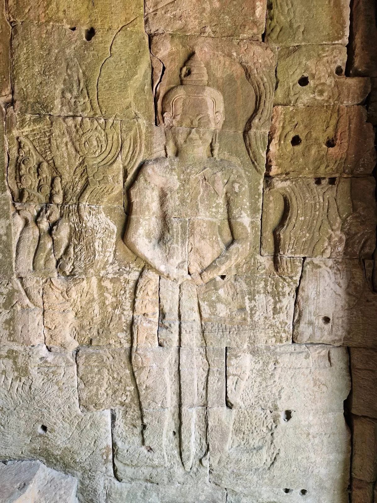 Ancient stone relief carving of a standing human figure on a textured wall, with visible time-worn details and some erosion in the surrounding stone.