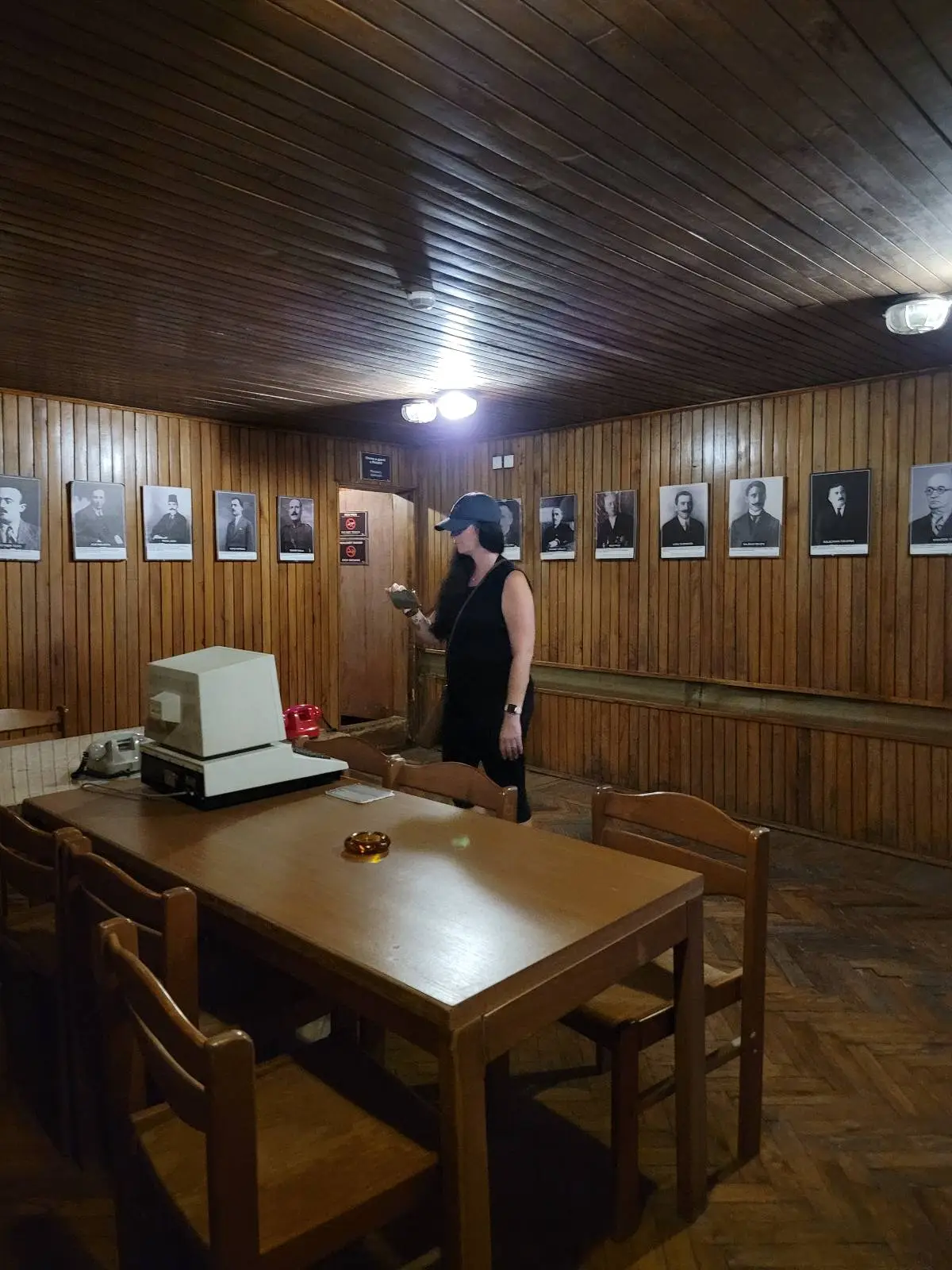 A person stands in a dimly lit, wood-paneled room with framed portraits on the walls. A table with an old computer and a wooden chair are in the foreground.