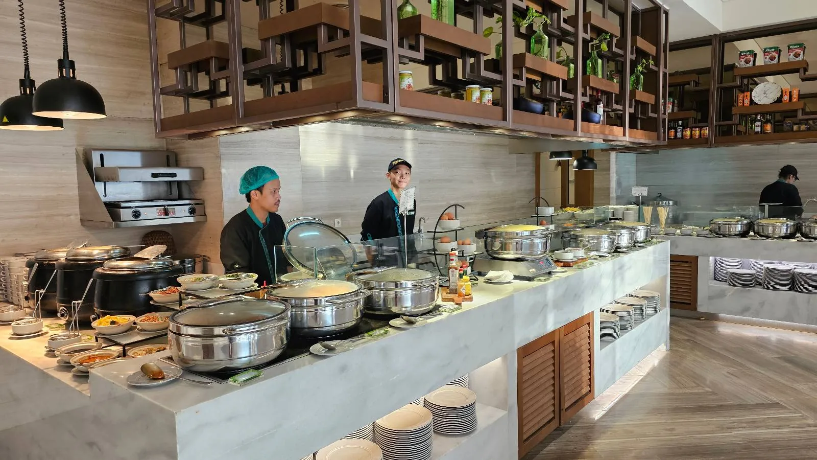 A buffet setup in a restaurant features various dishes in metal trays, with two staff members standing behind the counter. Plates are stacked at the front, and shelves with cups and bowls are visible above.