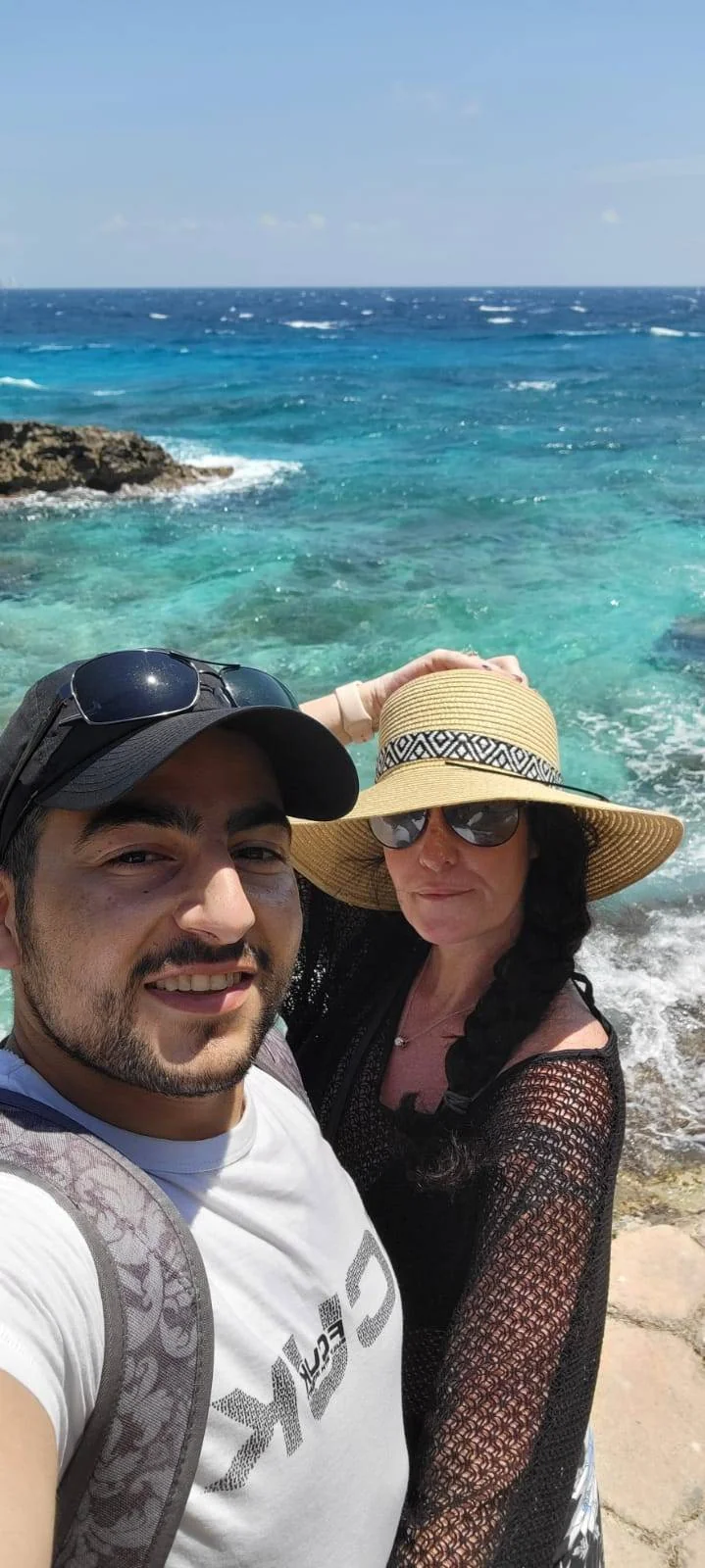 A couple takes a selfie on a sunny beach. The man is wearing a white t-shirt, cap, and backpack, while the woman is in a sun hat and black cover-up. Behind them, turquoise waves gently crash against rocky shorelines. The sky is clear and bright.