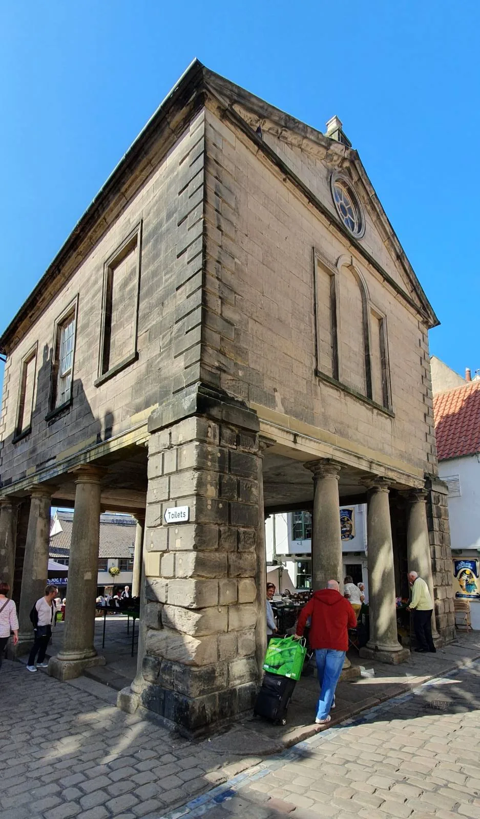 A tall, historical stone building with arched windows stands on pillars above a cobblestone street. People walk by, and the sky above is clear and blue.