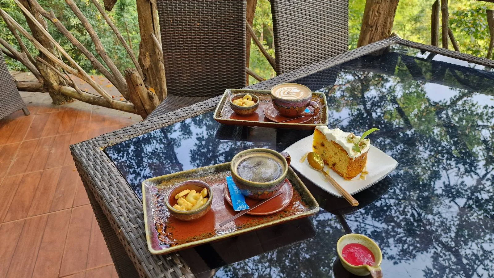 A glass-top table outdoors holds two trays with bowls of soup, bread, and condiments, a plate with a slice of cake, and a small cup of pink drink, surrounded by wicker chairs and greenery.