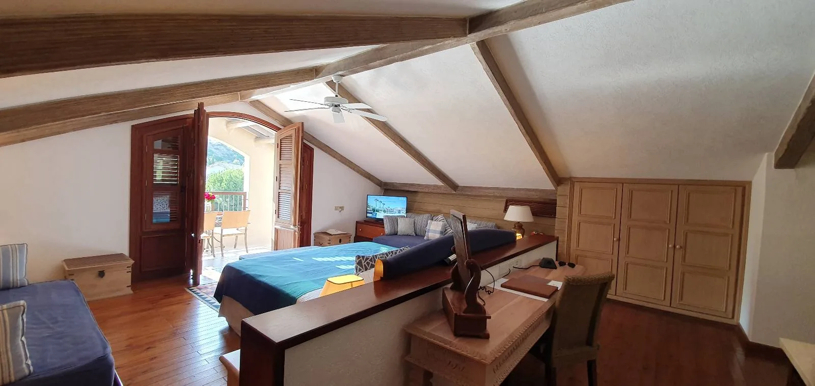 A cozy attic bedroom with a sloped ceiling features a double bed and single bed, wooden furniture, a small desk with a chair, and a TV. A door opens to a balcony with views, allowing natural light to fill the room.