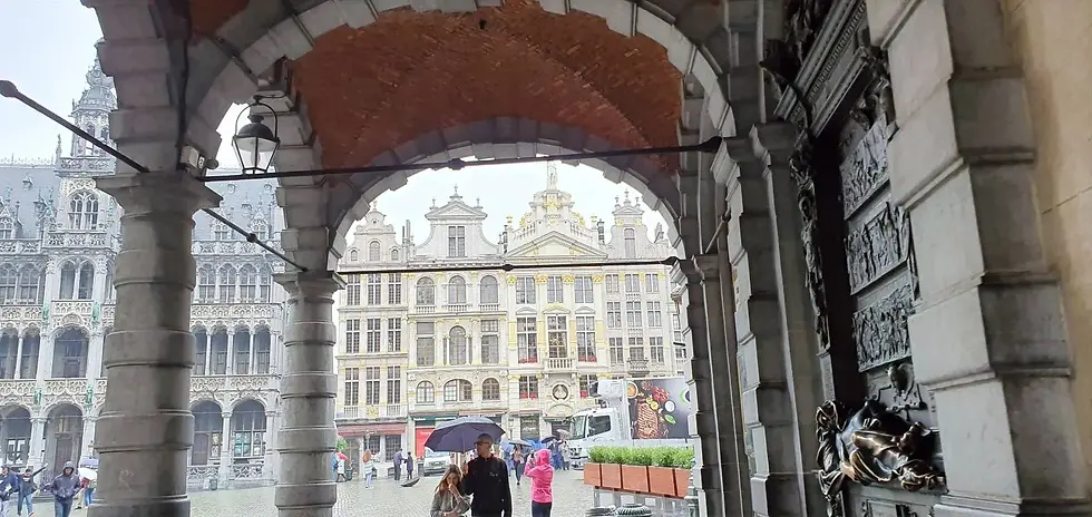 People with umbrellas walk through an archway, revealing ornate buildings with golden details in a cobblestone square. Overcast sky.