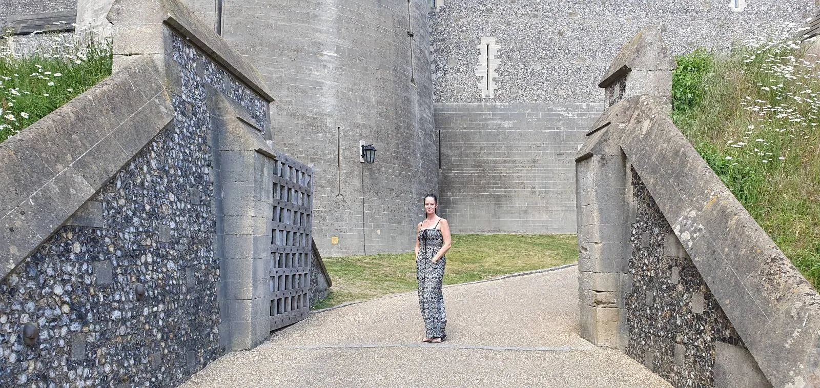 A person stands at the entrance of a stone castle, surrounded by large walls and an open metal gate. The pathway is bordered by greenery on one side and stone walls on the other.
