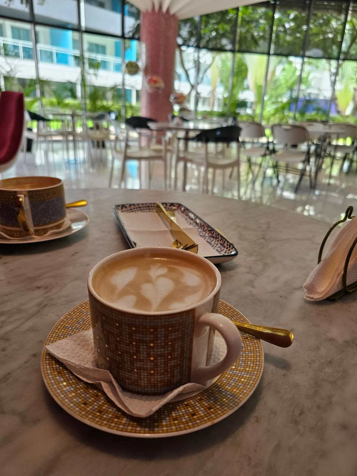 A coffee with milk froth sits on a patterned cup and saucer on a table. Another cup and a small tray with utensils are in the background. The setting is a bright cafe with large windows and modern decor.