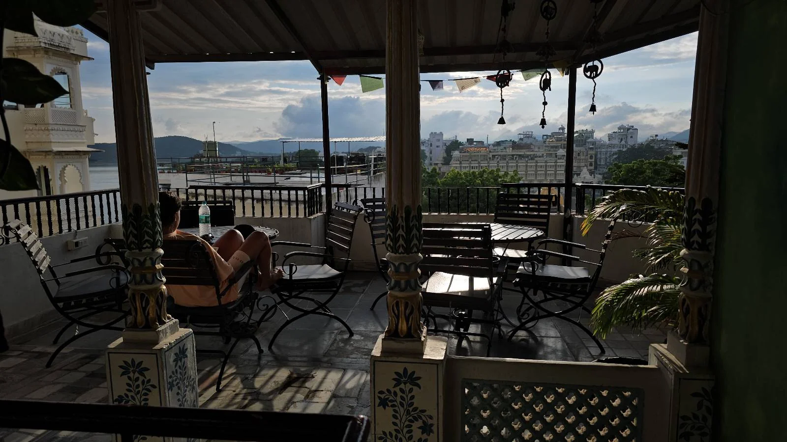 Outdoor terrace with metal chairs and tables, some people seated and relaxing. Hanging decorations and potted plants are visible, with a scenic city view and distant hills under a partly cloudy sky in the background.