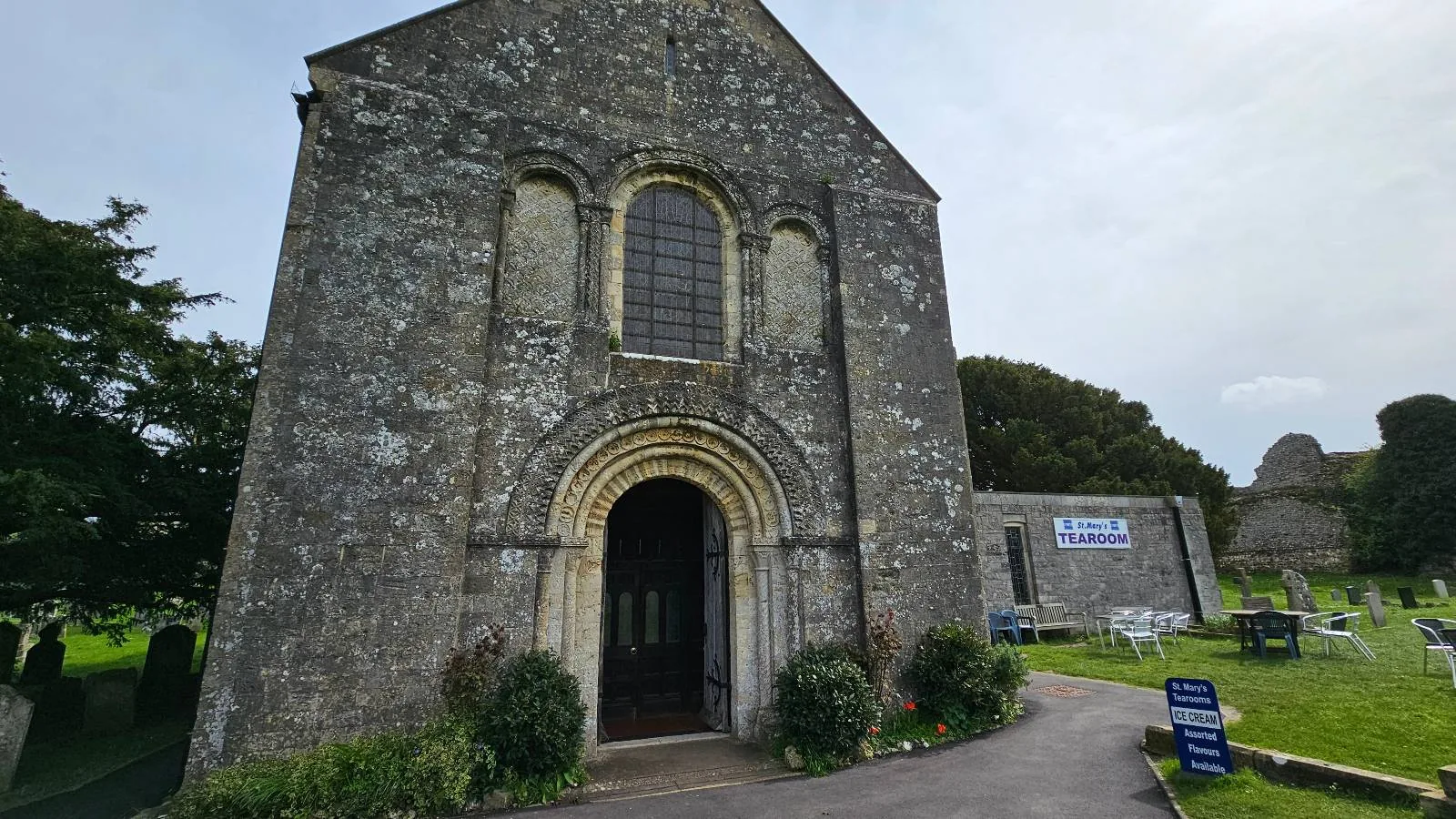Front view of an old stone church with an arched doorway and round windows, flanked by greenery and signs for a tearoom.