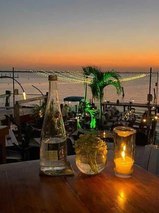 A sunset view over the ocean with a wooden table in the foreground. On the table, there's a candle, a glass of water, and a small plant. The background features palm trees, outdoor seating, and string lights.