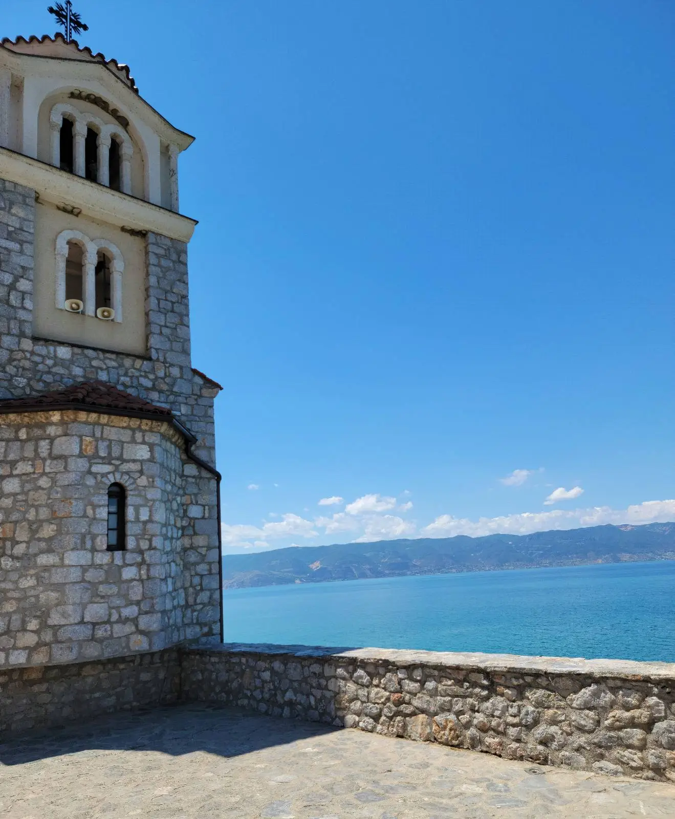 A stone building with an arched window and a cross on top stands in the foreground. In the background, a clear blue sky stretches above a calm, expansive body of water with distant hills. The scene is bright and serene.