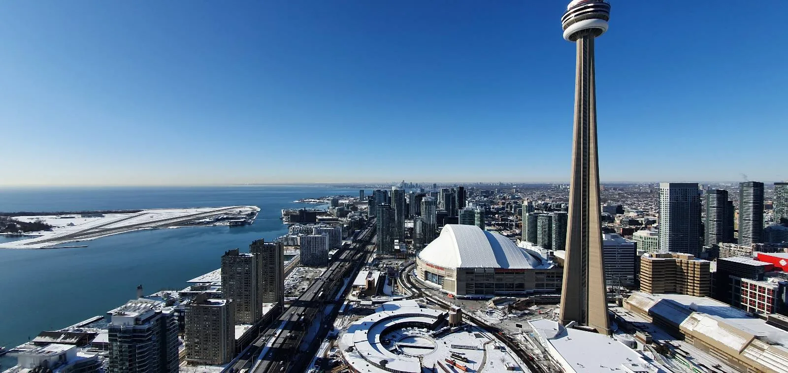 Aerial view of a cityscape with a prominent tall tower, a dome-shaped stadium, and high-rise buildings. A body of water lies to the left, bordered by snowy land. The sky is clear and blue.
