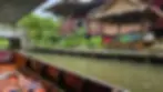 Boat on a canal beside wooden houses, lush greenery, and a riverside shop with drinks under a thatched roof. Calm and scenic setting.