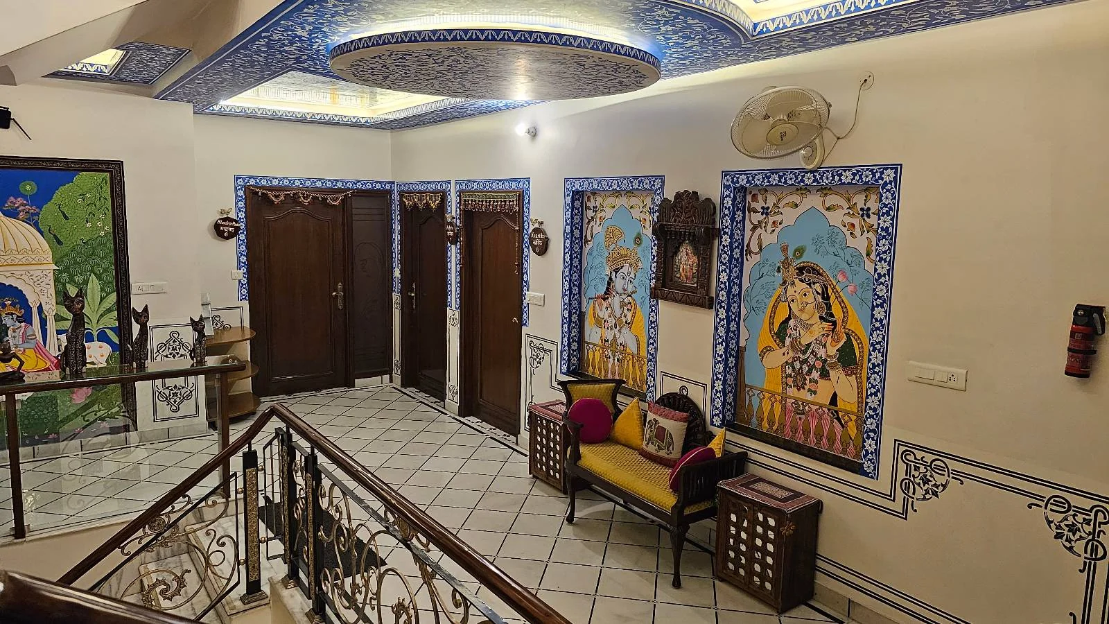 A hallway with tiled floors, wooden doors, vibrant wall art depicting traditional Indian scenes, a yellow cushioned bench with pillows, and decorative blue borders along the ceiling and walls.