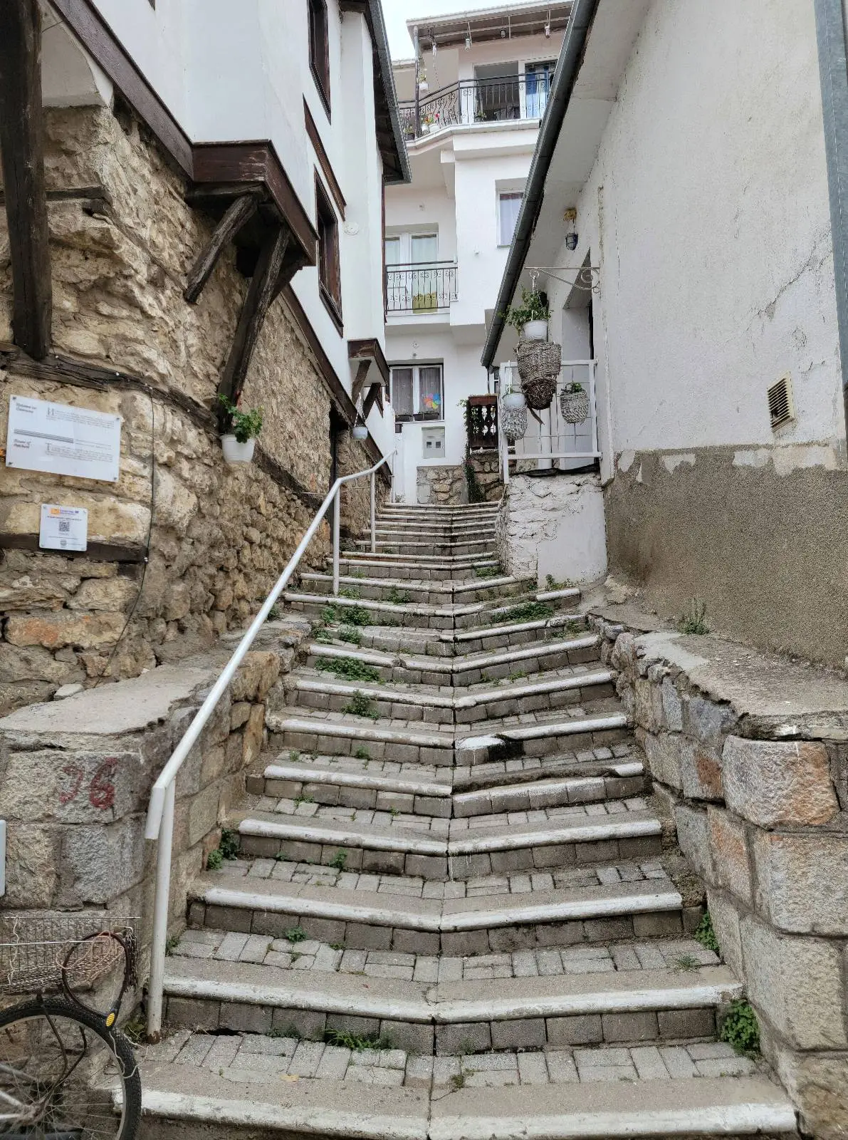 A narrow cobblestone stairway lined with stone and plastered buildings on either side, leading up to a higher level. A metal handrail runs along the left side, and some vegetation grows between the steps.