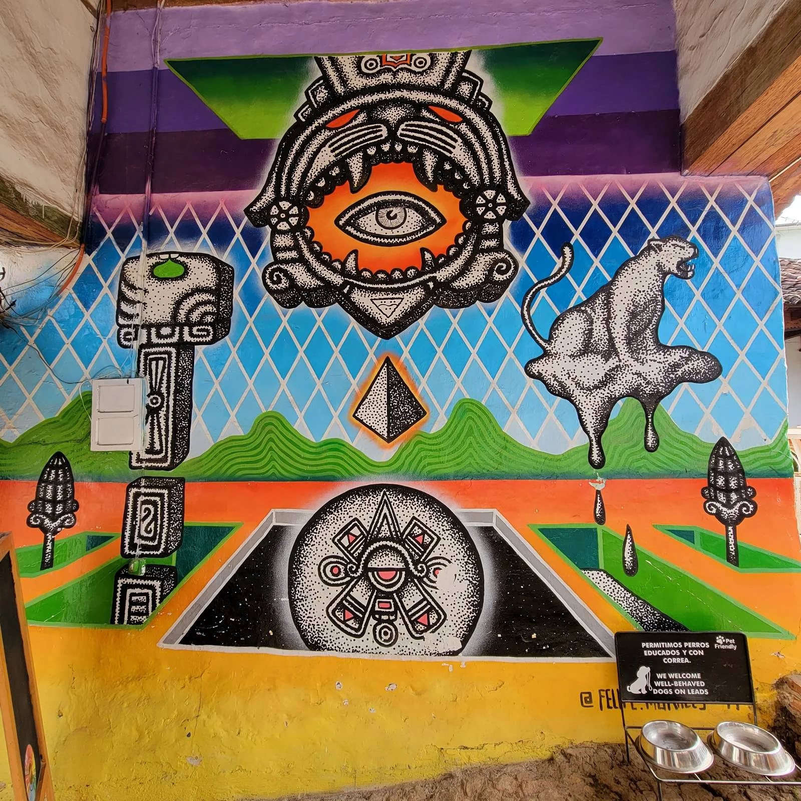 A vibrant mural featuring an eye within a tiger's mouth, geometric shapes, and a floating polar bear dripping liquid. The background is a colorful landscape of green fields and mountains under a blue sky with diamond patterns.